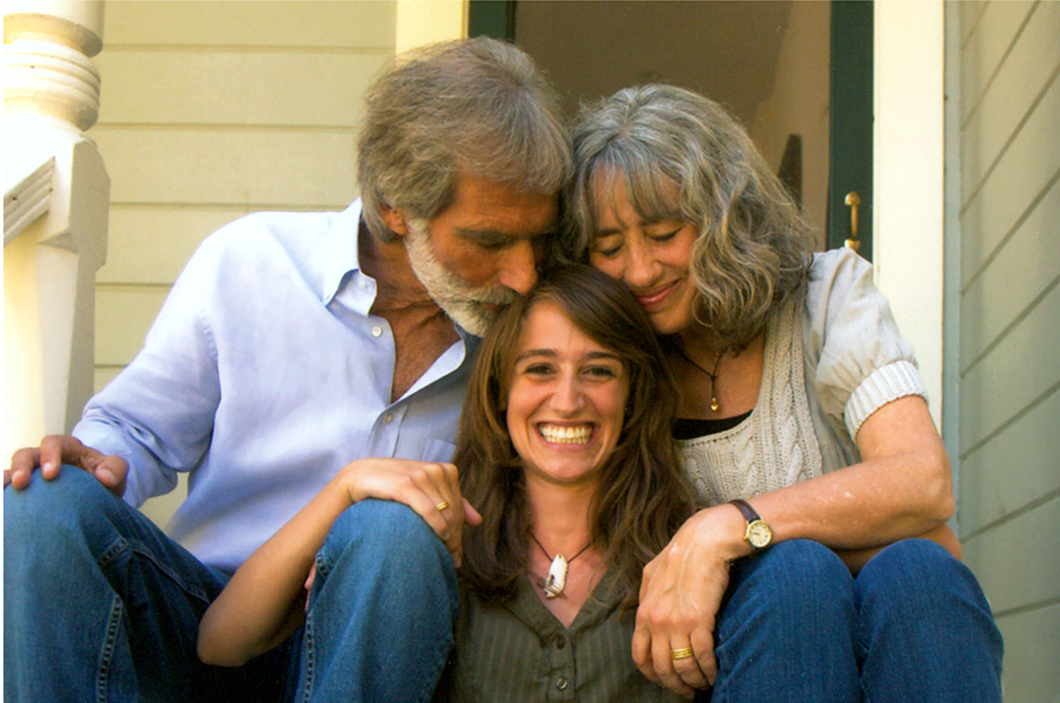 Smiling young woman sitting on porch steps embraced by two older adults, likely her parents.
